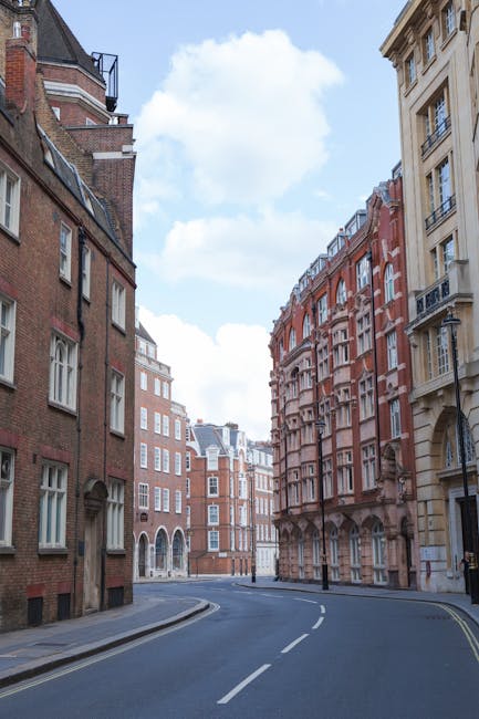 A wide view of a quiet residential street in a city, with red and beige multi-storey buildings featuring bay windows and decorative facades lining both sides of the road. The asphalt street curves gently to the left and is marked with white dashed lines. The sky is blue with scattered white clouds, and there are no vehicles or people visible. The scene depicts an urban environment suitable for house removals or furniture transport, with the calm street providing space for loading and unloading, which Knightsbridge Man and Van might utilise for home relocation services.