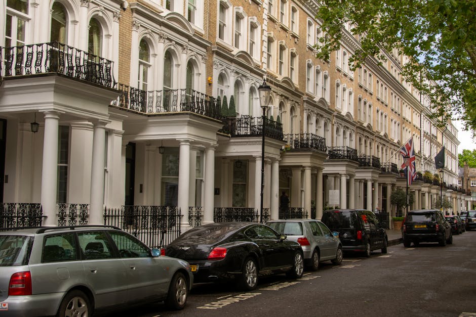 Photograph of a residential street in Knightsbridge with elegant, white-painted terraced houses featuring black wrought-iron balconies and decorative columns at the entrance. Several cars, including a silver estate, a black sports car, and other vehicles, are parked along the curb on a paved road with clear parking bays. The scene depicts a typical London neighbourhood, with a row of houses showing large windows, some with potted plants on balconies, and a lamppost visible in front of the buildings. Green tree foliage partially shades the street, and flags are hanging from some of the houses, adding to the characteristic appearance of the area. This image captures the environment where professional house removals, such as those provided by Knightsbridge Man and Van, might be involved in a home relocation or furniture transport process, with the loading area at the front of the properties ready for moving activities.